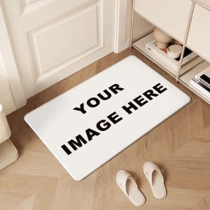 Puede incluir: Felpudo rectangular blanco con el texto "YOUR IMAGE HERE" en letras negras en negrita. Dos pares de zapatillas blancas están en el suelo de madera. La alfombra está colocada frente a una puerta blanca.