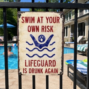 Może przedstawiać: Prostokątny metalowy znak z postarzanym, kremowobiałym wykończeniem, zawierający czerwony napis SWIM AT YOUR OWN RISK oraz LIFEGUARD IS DRUNK AGAIN, z centralną niebieską grafiką przedstawiającą osobę pływającą w falującej wodzie.