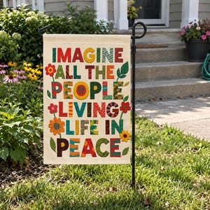 May include: A rectangular garden flag with a cream background featuring the text IMAGINE ALL THE PEOPLE LIVING LIFE IN PEACE in multi-colored, patterned block letters, decorated with various colorful flower illustrations and green leaves.