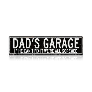 May include: A rectangular black metal sign with white text that reads "DAD'S GARAGE" above "IF HE CAN'T FIX IT WE'RE ALL SCREWED." The sign has a distressed, vintage look with a reflective surface.
