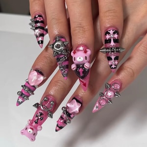 May include: A set of long, pointed acrylic nails with a pink and black color scheme. The nails feature various embellishments, including metal spikes, stars, hearts, and a cartoon bear. The designs incorporate stripes, crosses, and glitter.
