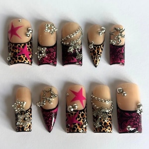 May include: A collection of ten artificial nails with a nude base, showcasing diverse designs. Some nails feature a pink star, others have silver rhinestones, and some display a leopard print pattern with a pink lace design. The nails are adorned with silver chain and rhinestone accents.