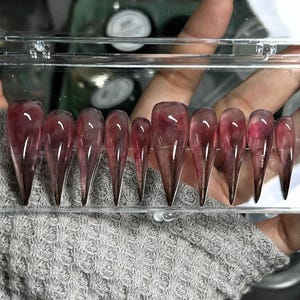 Puede incluir: Un conjunto de diez uñas postizas largas y puntiagudas, transparentes. Las uñas son de color burdeos oscuro con un efecto veteado. Están dispuestas en una caja de plástico transparente.