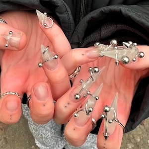 May include: A hand with long, clear acrylic nails adorned with silver-colored jewelry. The nails feature silver barbells, rings, and spikes. Some nails have a clear, pointed shape, while others have a flat, rectangular shape.