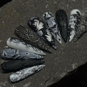 May include: A collection of nine stiletto-shaped press-on nails featuring intricate 3D designs in black, white, and gray. The set includes various textures, including smooth matte finishes, glossy raised patterns, marbled effects, and metallic silver accents.