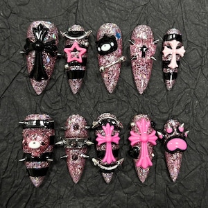 May include: A set of ten press-on nails with a pink glitter base and black horizontal stripes. The nails feature 3D embellishments including black and pink crosses, a black bear face, a pink star, silver metal studs, and a pink paw print design.
