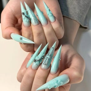 Op de afbeelding: Stiletto-vormige nagels met een licht mintgroene ombre-afwerking. De nagels zijn voorzien van driedimensionale zilveren kruisbedels, metalen klinknagels en gedetailleerde zilveren lijnen. Het ontwerp bevat een kruispatroon omlijnd met kleine zilveren kraaltjes.