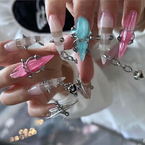 May include: Long, pointed artificial nails featuring a translucent finish in shades of pink, blue, and clear. Each nail is adorned with silver-toned metal hardware, including small spikes, circular rings, and a delicate chain with a heart charm.
