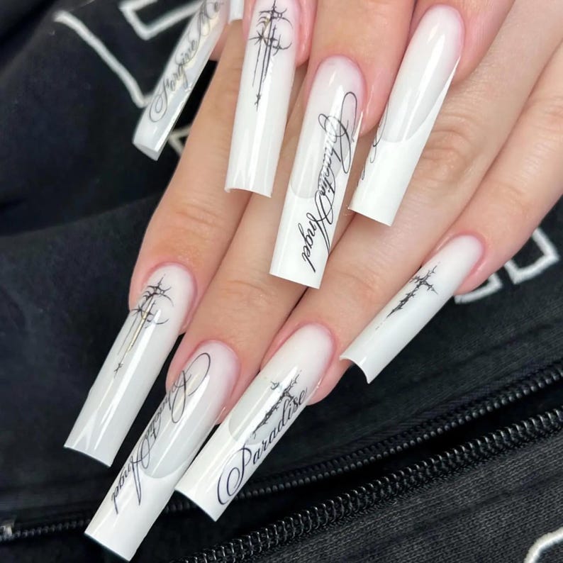 Pode incluir: Unhas compridas de acr&iacute;lico branco com acabamento brilhante. Cada unha apresenta escrita preta e desenhos gr&aacute;ficos, incluindo as palavras "Paradise" e "Angel". As unhas t&ecirc;m formato quadrado e um gradiente sutil na base.