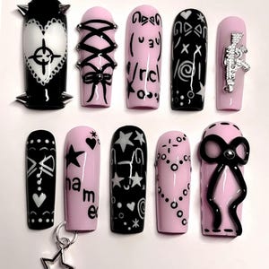 Könnte beinhalten: Zehn künstliche Fingernägel in Schwarz und Rosa mit verschiedenen Designs. Die Designs umfassen Schleifen, Sterne und Text, einige mit silbernen Metallakzenten. Ein Nagel hat einen Sternanhänger.