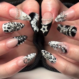 Puede incluir: Un conjunto de uñas manicuradas con varios diseños. Algunas uñas son negras y blancas, con patrones como estampado de vaca y rayas de cebra. Otras están adornadas con cadenas plateadas, tachuelas y adornos en forma de estrella. Las uñas tienen un acabado brillante.