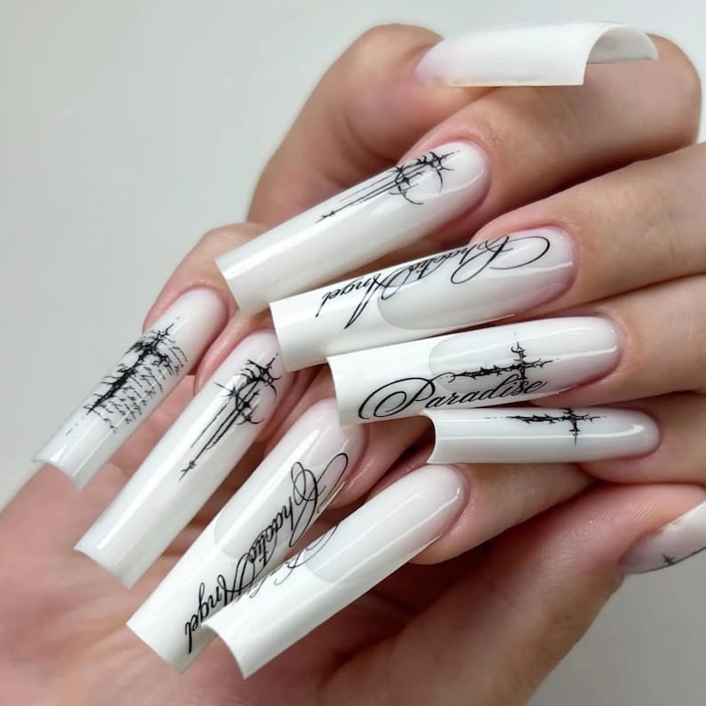Pode incluir: Unhas acr&iacute;licas longas e brancas com desenhos e texto pretos em estilo g&oacute;tico. O texto inclui as palavras "Chaos Angel" e "Paradise". As unhas t&ecirc;m um formato quadrado e alongado.