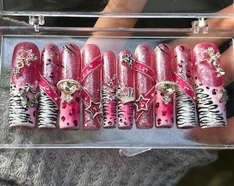 May include: A set of ten press-on nails in a clear plastic case. The nails feature a pink glitter base with various designs, including zebra stripes, leopard spots, and rhinestone embellishments. Some nails have star and crown accents.