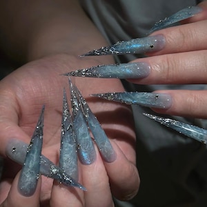 Puede incluir: Uñas artificiales largas en forma de estilete con un efecto degradado marmoleado en azul y gris. La superficie está decorada con intrincados detalles metálicos plateados y pequeños cristales transparentes incrustados, creando un aspecto gélido.