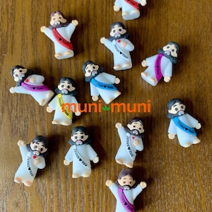 May include: A collection of small, white figurines depicting a figure with a beard and long hair, wearing a white robe. Each figurine has a sash with the text "Jesus You" in various colors. The figurines are arranged on a wooden surface.