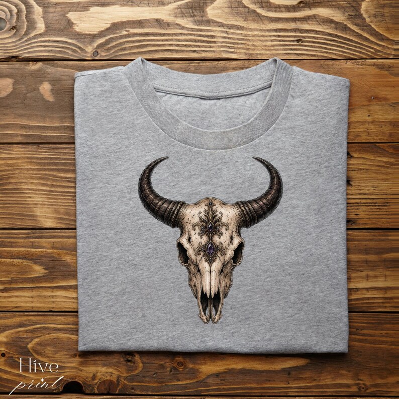 May include: A heather grey crew neck t-shirt featuring a central graphic of a detailed animal skull with large curved horns. The skull is decorated with intricate filigree patterns and two dark purple gemstone accents centred on the forehead.