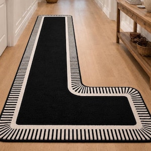 Op de afbeelding: Een L-vormig tapijt met een zwart middenstuk en een witte rand met zwarte strepen. Het tapijt ligt op een houten vloer. Het ontwerp is een modern, geometrisch patroon.