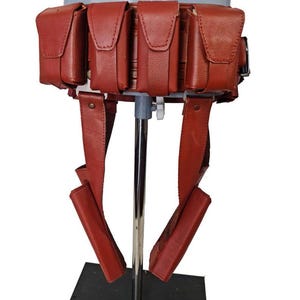 May include: A red leather utility belt with multiple pouches and straps. The belt is designed to be worn around the waist and features several rectangular pouches with flap closures. The straps hang down from the belt.