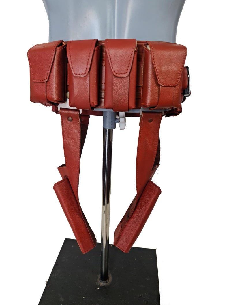 May include: A red leather utility belt with multiple pouches and straps. The belt is designed to be worn around the waist and features several rectangular pouches with flap closures. The straps hang down from the belt.
