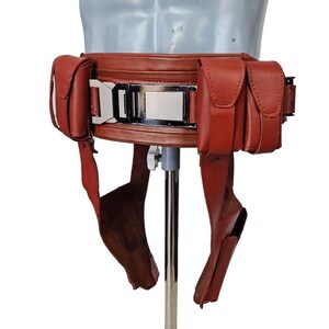 May include: A reddish-brown leather belt with multiple pouches and a silver buckle. The belt has two pouches on each side and two straps hanging down. The belt is displayed on a mannequin.