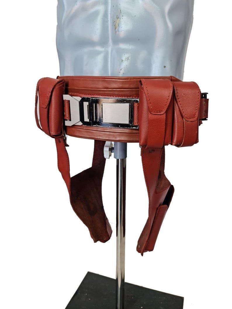 May include: A reddish-brown leather belt with multiple pouches and a silver buckle. The belt has two pouches on each side and two straps hanging down. The belt is displayed on a mannequin.