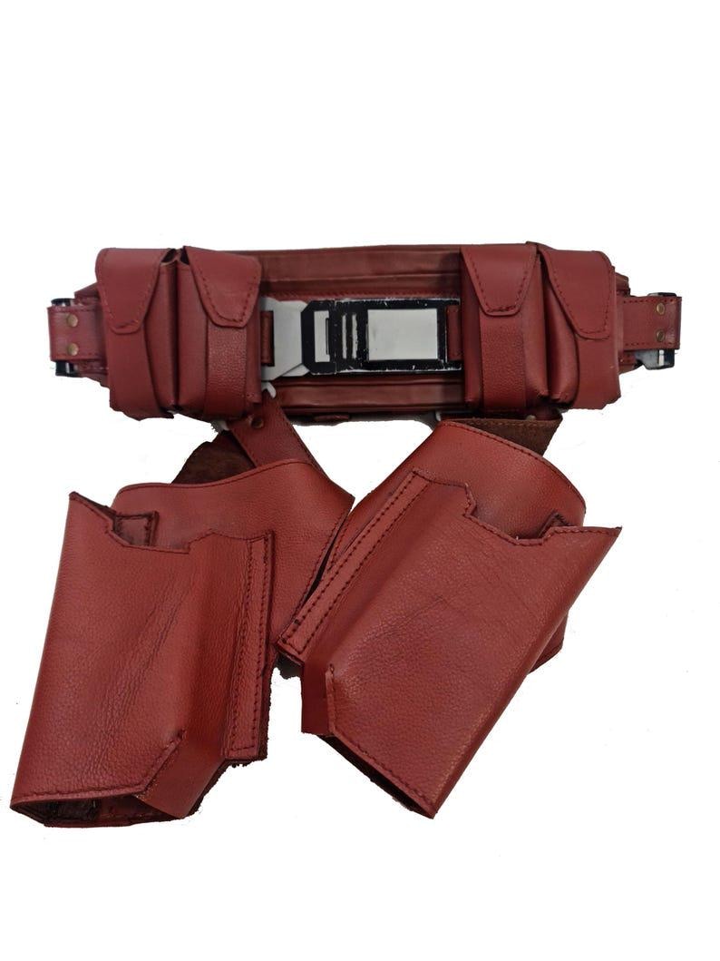 May include: A burgundy leather utility belt with multiple pouches and a rectangular buckle. The belt features several small, square pouches and two larger holster-like pouches. The leather appears to be stitched with a darker thread.