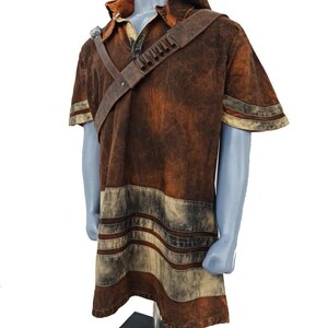 May include: A short-sleeved hooded tunic in a distressed brown finish with a weathered texture. The garment features a diagonal shoulder strap with small cylindrical loops and horizontal light-colored bands near the hem and sleeves.