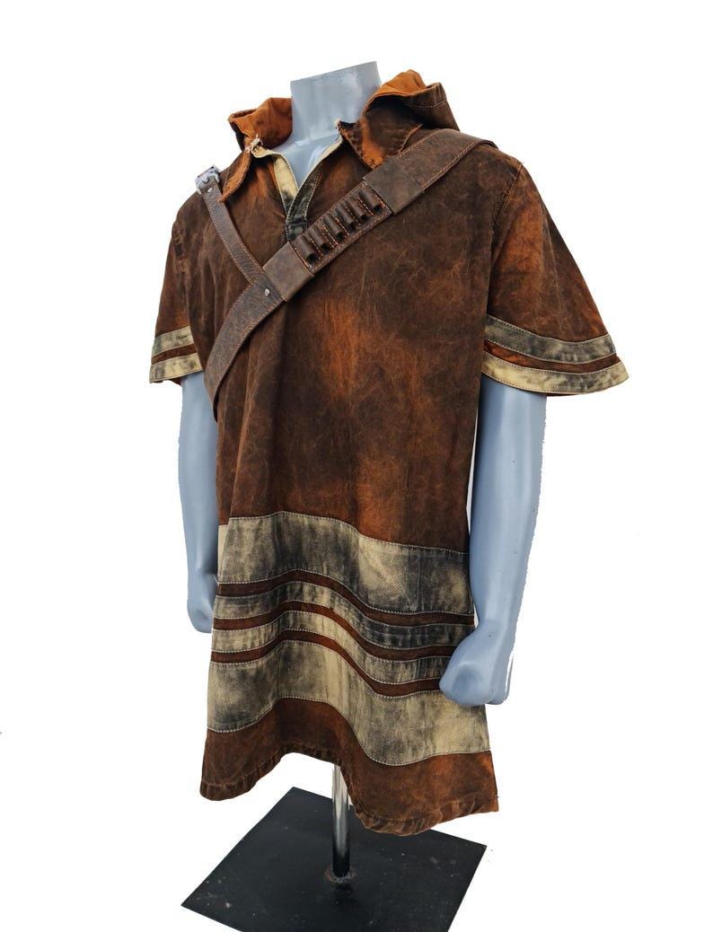 May include: A short-sleeved hooded tunic in a distressed brown finish with a weathered texture. The garment features a diagonal shoulder strap with small cylindrical loops and horizontal light-colored bands near the hem and sleeves.