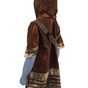 May include: A short-sleeved hooded tunic in a distressed brown and tan wash. The garment features a diagonal strap across the chest with a black metal buckle and multiple horizontal bands of lighter tan fabric along the hem and sleeves.