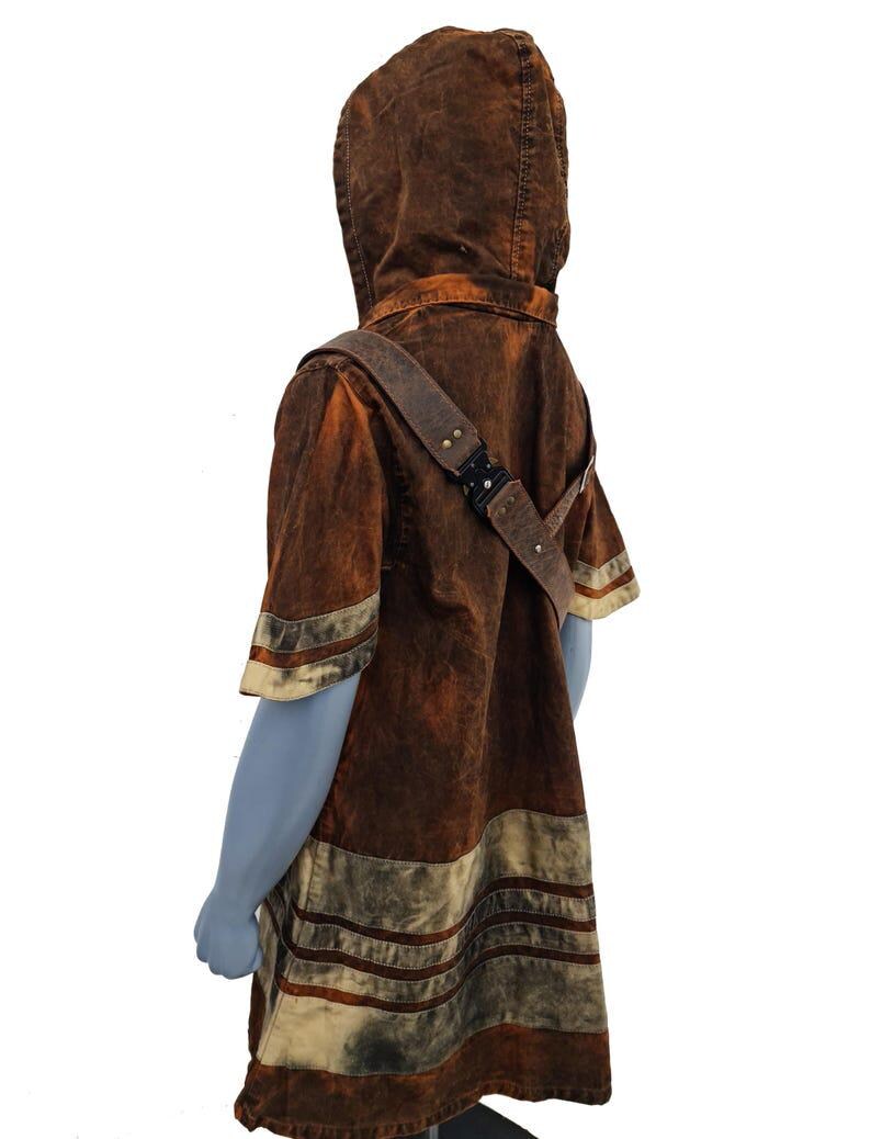 May include: A short-sleeved hooded tunic in a distressed brown and tan wash. The garment features a diagonal strap across the chest with a black metal buckle and multiple horizontal bands of lighter tan fabric along the hem and sleeves.