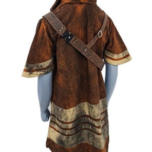 May include: A short-sleeved hooded tunic in a distressed brown finish with lighter beige horizontal bands at the hem and sleeves. A brown leather-like strap with a black metal buckle crosses the chest diagonally over the garment.