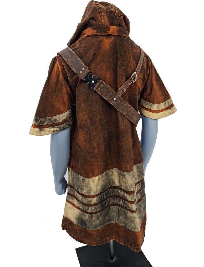 May include: A short-sleeved hooded tunic in a distressed brown finish with lighter beige horizontal bands at the hem and sleeves. A brown leather-like strap with a black metal buckle crosses the chest diagonally over the garment.