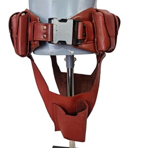May include: A red leather utility belt with pouches and a buckle. The belt has two pouches on each side and a central buckle. A lower section with a pocket hangs down from the belt.