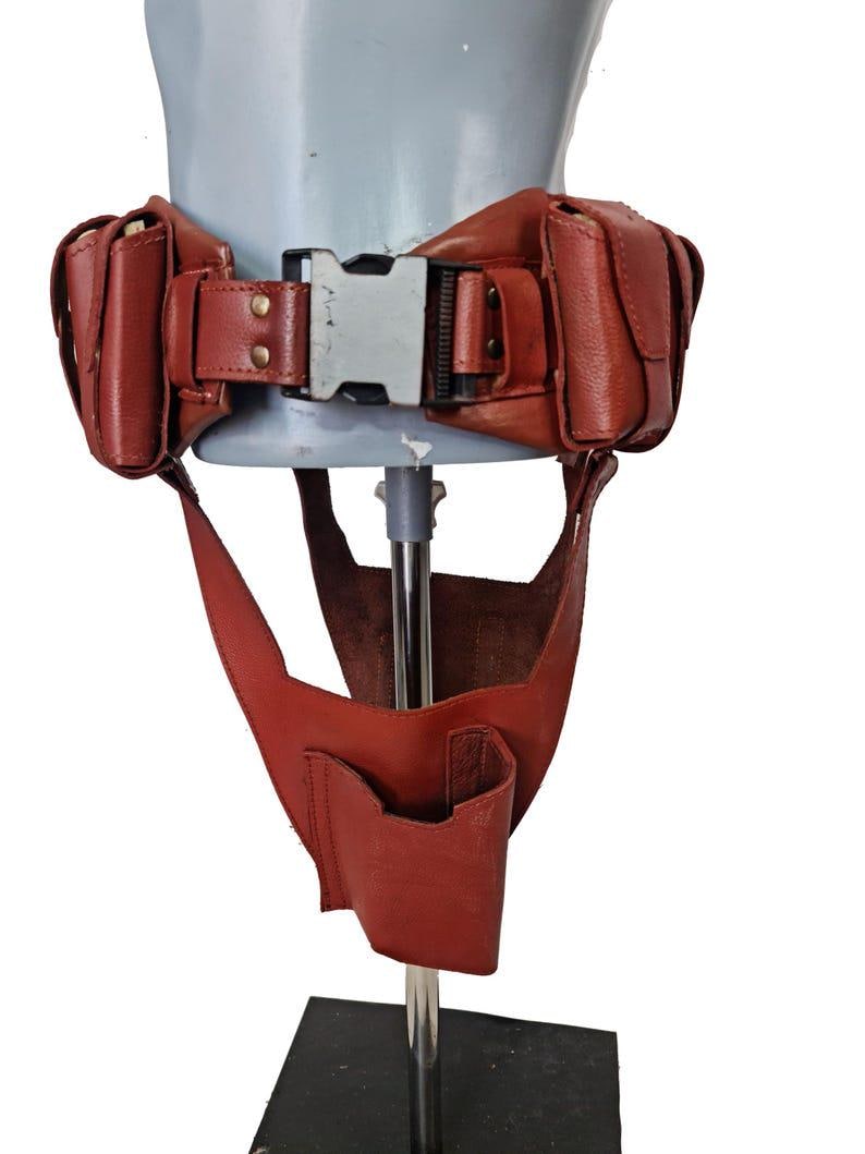 May include: A red leather utility belt with pouches and a buckle. The belt has two pouches on each side and a central buckle. A lower section with a pocket hangs down from the belt.