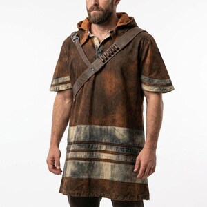 May include: A short-sleeved hooded tunic in a distressed brown finish with a weathered texture. The garment features horizontal light-colored bands near the hem and sleeves, and includes a brown leather-like shoulder strap with metal hardware.