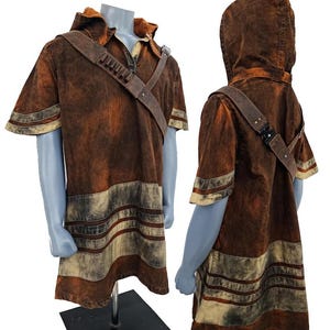 May include: A short-sleeved hooded tunic in a distressed brown and tan wash. The garment features a diagonal shoulder strap with small cylindrical attachments and a buckle. The lower section displays horizontal bands of alternating brown and tan colors.