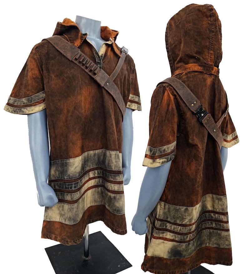 May include: A short-sleeved hooded tunic in a distressed brown and tan wash. The garment features a diagonal shoulder strap with small cylindrical attachments and a buckle. The lower section displays horizontal bands of alternating brown and tan colors.