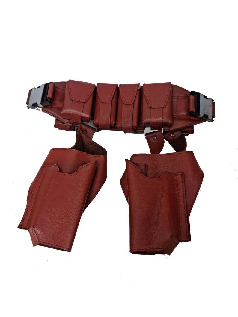 May include: A brown leather utility belt with multiple pouches and pockets. The belt features a silver buckle and two additional pouches hanging below. The design is symmetrical and appears to be made of high-quality leather.