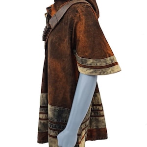 May include: A hooded tunic in a mottled brown and rust-colored finish, featuring short sleeves with light-colored horizontal bands. A dark brown strap with a series of small, cylindrical attachments is worn diagonally across the chest area.
