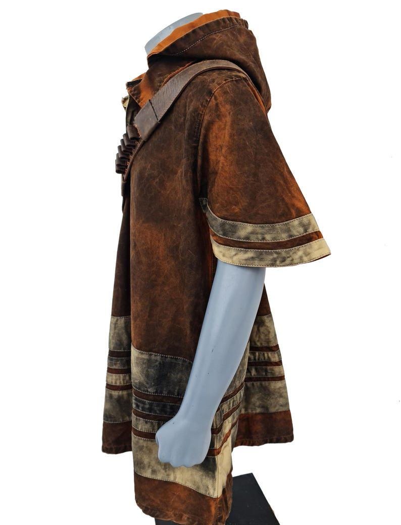 May include: A hooded tunic in a mottled brown and rust-colored finish, featuring short sleeves with light-colored horizontal bands. A dark brown strap with a series of small, cylindrical attachments is worn diagonally across the chest area.