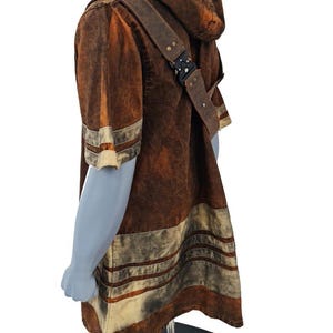 May include: A brown hooded short-sleeve tunic with a distressed, mottled texture. The garment features horizontal cream-colored stripes on the sleeves and hem, and includes a brown shoulder strap secured with a black metal buckle mechanism.