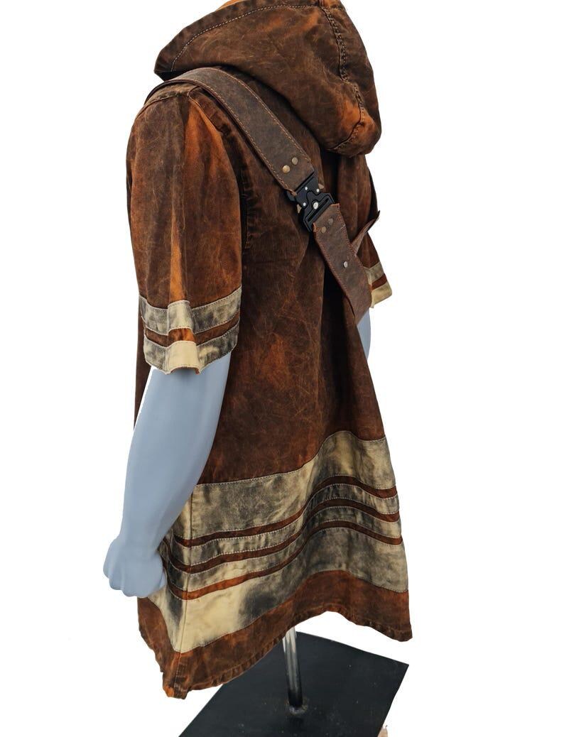 May include: A brown hooded short-sleeve tunic with a distressed, mottled texture. The garment features horizontal cream-colored stripes on the sleeves and hem, and includes a brown shoulder strap secured with a black metal buckle mechanism.
