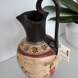 Puede incluir: Jarra de cerámica con asa y borde marrón oscuro, con cuerpo beige con figuras pintadas y texto griego. La jarra tiene una banda decorativa y una etiqueta que dice "SOBONI CERAMICS".