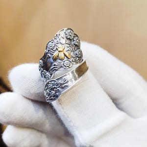Pode incluir: Anel prateado com uma abelha dourada e design floral. O anel tem um estilo envolvente e é adornado com pequenos detalhes brilhantes. A abelha está centrada, adicionando um toque de elegância inspirado na natureza.