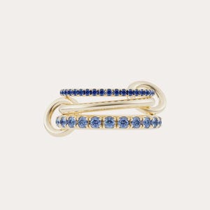 May include: Three-ring gold jewelry set. The top and bottom rings are adorned with small blue gemstones. The rings are linked together with a gold connector. The jewelry is set against a plain white background.