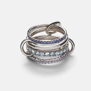 May include: A stack of silver rings with various designs. One ring features a knot design, while others are adorned with blue, light blue, and purple gemstones. The rings are connected by silver loops, creating a unique and stylish look.
