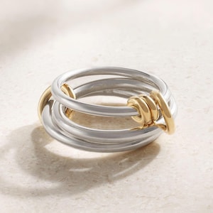 May include: A silver and gold ring with a unique interlocking design. The ring features three silver bands intertwined with gold accents. The gold elements are clustered together, creating a striking contrast.