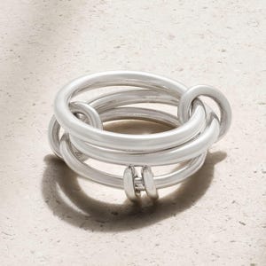 Peut inclure: Une bague argentée au design unique. La bague présente plusieurs bandes entrelacées, créant un aspect superposé et texturé. Le métal a une finition lisse et polie, reflétant la lumière et ajoutant à son esthétique moderne.