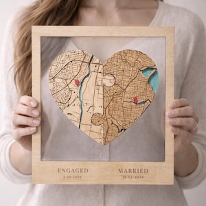 May include: A framed wooden heart-shaped map, featuring two puzzle pieces with red pin markers. The frame is light-coloured wood. The text "ENGAGED 2-11-2021" and "MARRIED 11-02-2026" are printed below the map.