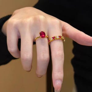 May include: Two gold rings with red gemstones. One ring features a large oval-cut ruby, while the other is an eternity band with alternating red and clear stones. The rings are set on a hand.
